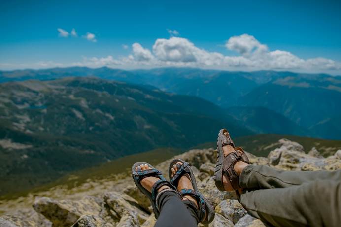 Angestrebtes Ziel der Ehe- und Paartherapie, egal, ob gemeinsame oder getrennte Wege: Ein liegendes Paar, das seine Füße entspannt gekreuzt hat, mit Blick auf ein Bergpanorama, blauem Himmel und Wolken.