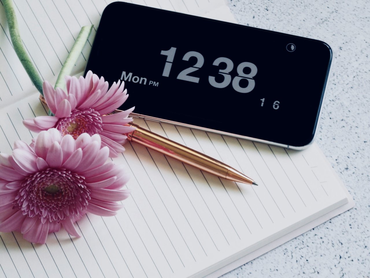 Foto von Jess Bailey auf Unsplash Offenes Schreibheft mit Smartphone, Stift und Blumen.